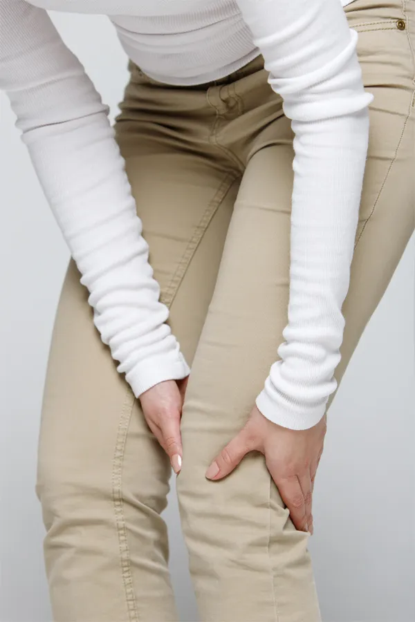 Arthritis-Treatment A woman wearing a white long-sleeve shirt and tan pants, clutching her knee, suffering from arthritis. Get arthritis treatment from Proactive Wellness Centers in Loudoun County.