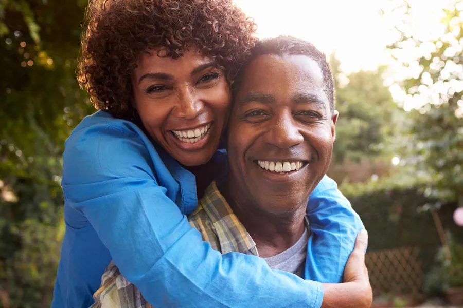 Autoimmune-Doctor A middle-aged black couple outside, the woman embracing the man from behind pressing her cheek to his, both are smiling due to successful Autoimmune Disorder treatment from Proactive Wellness Centers in Loudoun County.