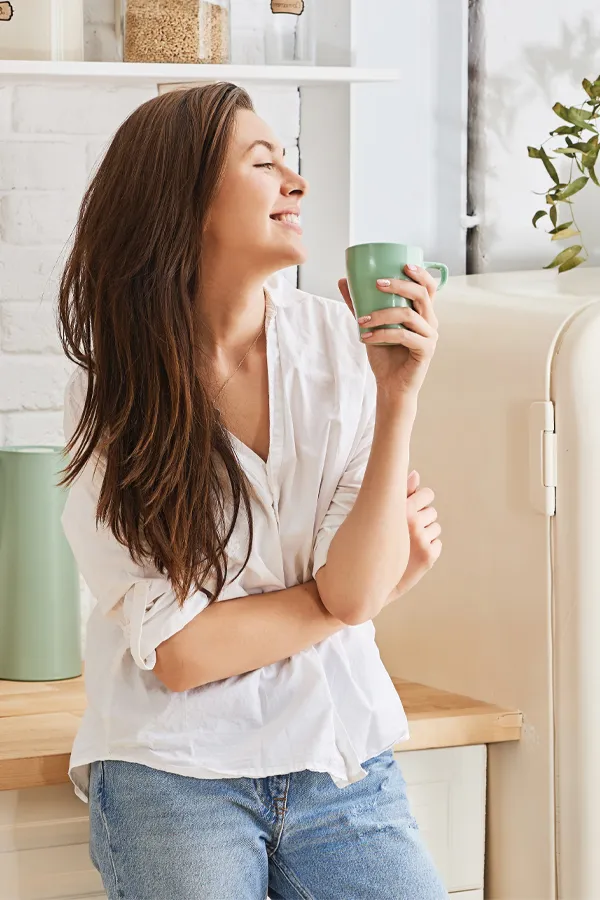 Crohns-Disease-Treatment A woman with a white blouse an jeans drinking tea in her kitchen, smiling after getting treatment for Crohn's Disease from Proactive Wellness Centers in Loudoun County.