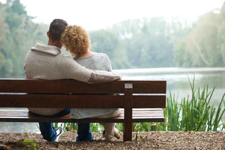 A couple wearing sweaters seen from behind sitting on a bench and looking out at a relaxing lake and forest, representing functional medicine treatment from Proactive Wellness Centers in Loudoun County.
