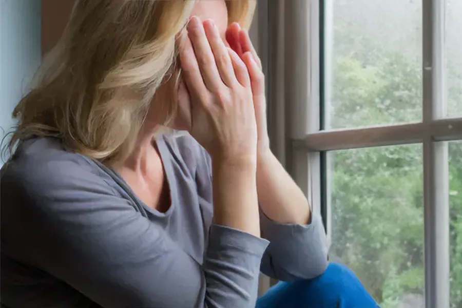 Graves-Disease-Clinic A blonde woman in a gray shirt sits in front of a window on a rainy day covering her eyes with her hands. Get treatment for Grave's Disease from Proactive Wellness Centers in Loudoun County.
