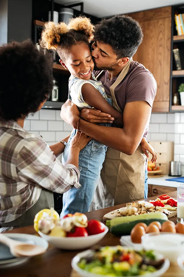 Gut-Health-Treatment A man holds his daughter and kisses her cheek as the family makes a healthy dinner in the kitchen. Get help for digestive issues and gut health from Proactive Wellness Centers in Loudoun County.