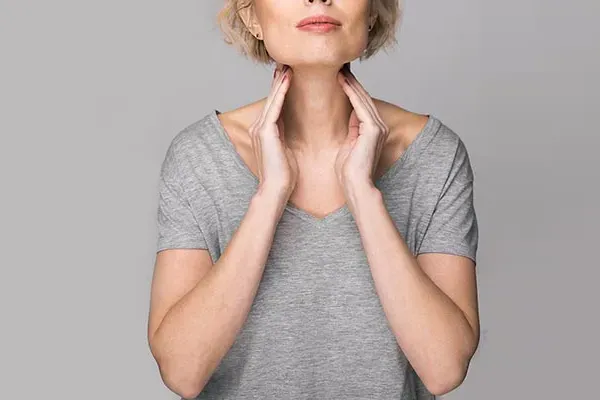 Hashimotos-Thyroiditis-Doctor An older blonde woman in a gray t-shirt holds her hands to her neck checking for signs of thyroid problems. Get help for Hashimoto's Thyroiditis from Proactive Wellness Centers in Loudoun County.
