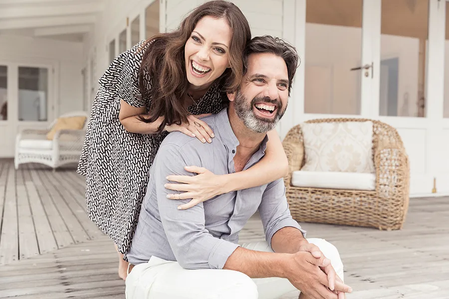 Health-and-Wellness-Services-Doctor A middle-aged couple on a wooden porch with wicker furniture; the woman is playfully hugging the man from behind. They are benefiting from the health and wellness services from Proactive Wellness Centers in Loudoun County.