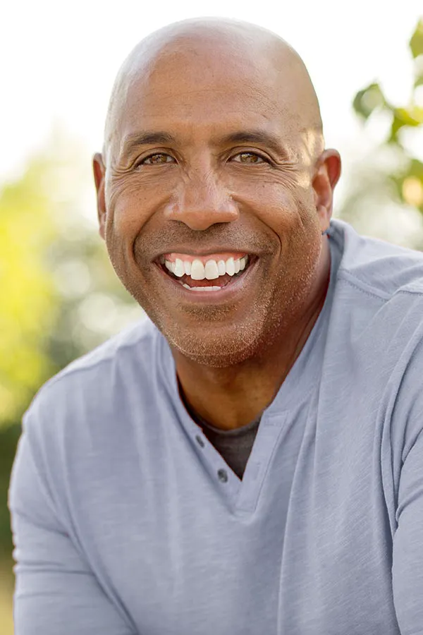 Healthy-Aging-Peptides-Treatment A middle-aged black man in a gray v-neck t-shirt sits outside smiling, happy with the healthy aging peptide treatments from Proactive Wellness Centers in Loudoun County