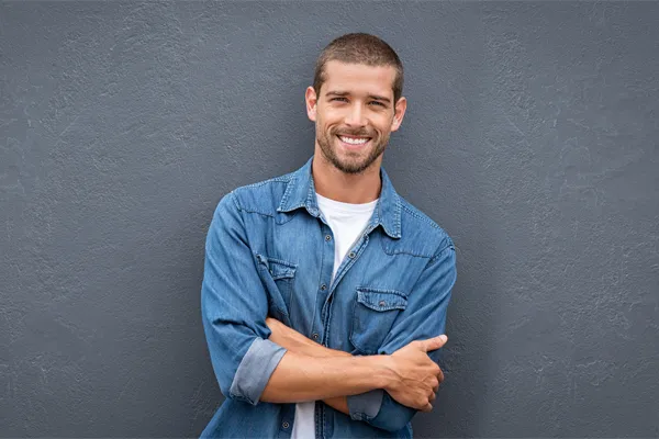 Hormones-for-Men-Doctor A man in a denim shirt stands smiling against a gray-blue wall, pleased with his testosterone hormone therapy from Proactive Wellness Centers in Loudoun County.