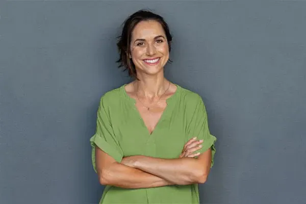 Hormones-for-Women-Doctor A woman in a green shirt stands smiling against a gray-blue wall, pleased with her hormone therapy from Proactive Wellness Centers in Loudoun County.