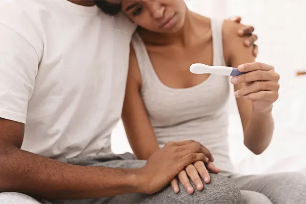 Infertility-Clinic A black couple sits on a bed, disappointed with the negative results of a pregnancy test. Get treatment for infertility from Proactive Wellness Centers in Loudoun County.