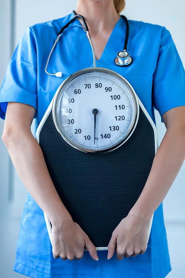 Medical-Weight-Loss-Treatment A nurse in blue scrubs with a stethoscope hanging around her neck holds a scale, representing weight loss treatments from Proactive Wellness Centers in Loudoun County.