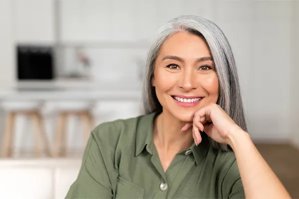 Menopause-Clinic An asian woman with long gray hair and a green shirt sitting and smiling on her couch, happy with her menopause care from [NAME} in Loudoun County.