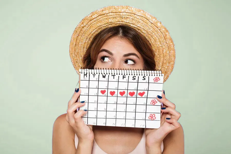 Menstrual-Dysfunction-Doctor A woman in a straw hat holds a calendar with several days marked off in front of her nose and mouth. Get treatment for menstrual dysfunction from Proactive Wellness Centers in Loudoun County.