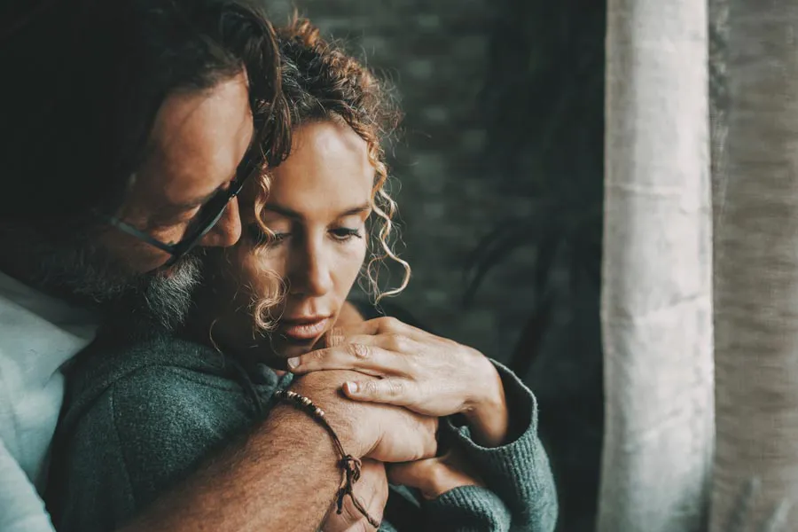 Neurodegenerative-Disorders-Therapy A man embraces a woman from behind in a dimly lit room, their somber expressions showing concerns about a neurodegenerative disease they will get treated by Proactive Wellness Centers in Loudoun County.