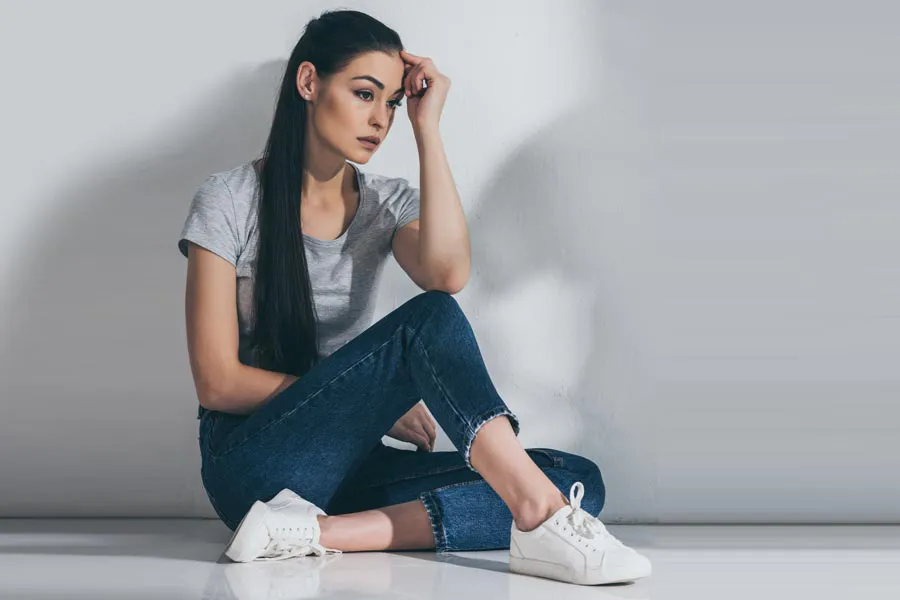 PCOS-Doctor A young brunette woman in a gray t-shirt and jeans sits against a gray wall with her head resting on her hand and a distressed expression. Get help for Polycystic Ovarian Syndrome (PCOS) from Proactive Wellness Centers in Loudoun County.