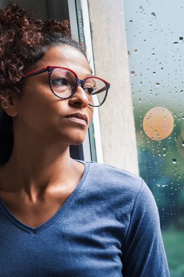 PMDD-Treatment A black woman with with a somber expression and glasses and a gray shirt stares out the window on a rainy day. Get treatment for Premenstrual Dysphoric Disorder from Proactive Wellness Centers in Loudoun County.