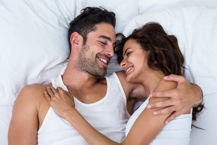 PT141-for-Men-Doctor A laughing man and woman in undershirts lie in each other's arms on a white bed, representing successful PT141 sexual wellness therapy for men from Proactive Wellness Centers in Loudoun County.