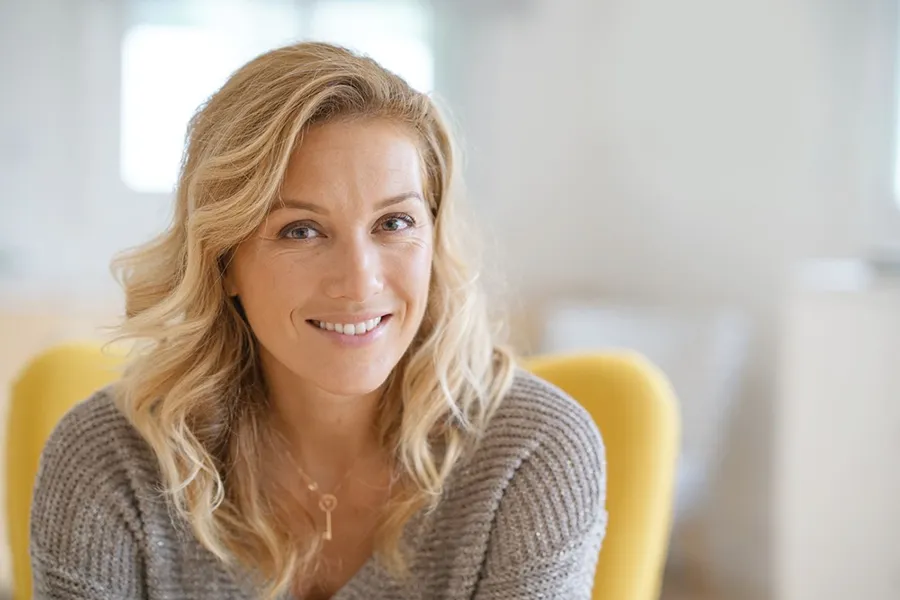 Perimenopause-Doctor A blonde woman wearing a a gray sweater sits inside in a yellow chair, smiling and happy with her perimenopause treatment from Proactive Wellness Centers in Loudoun County.