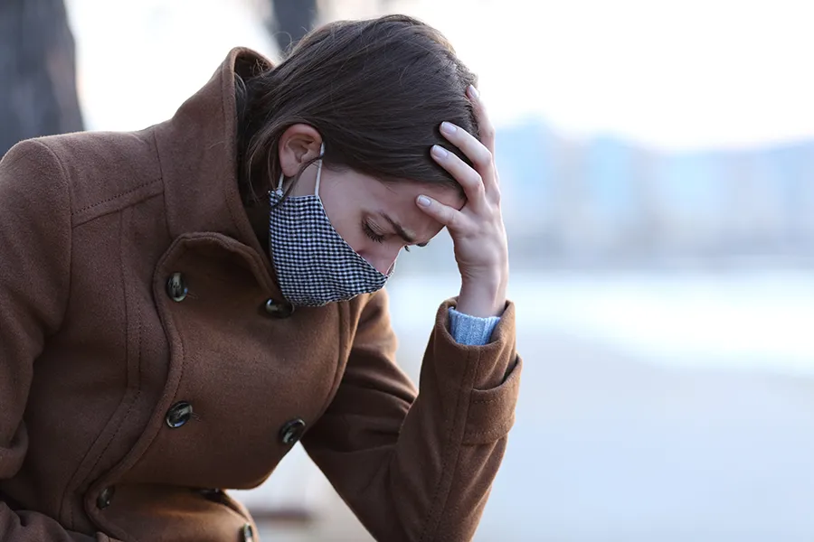 Post-Covid-Clinic A woman in a mask sits outside with her hand on her forehead in pain. Get post-covid treatment from Proactive Wellness Centers in Loudoun County.