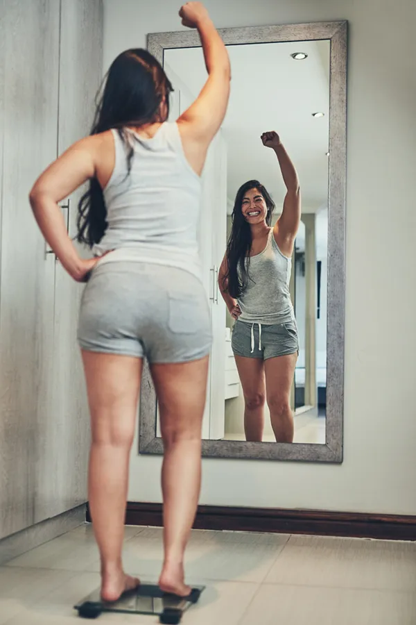 Tirzepatide-Treatment A woman in workout clothes stands on a scale looking in the mirror and pumping her hands in celebration of her successful Tirzepatide treatment from Proactive Wellness Centers in Loudoun County.